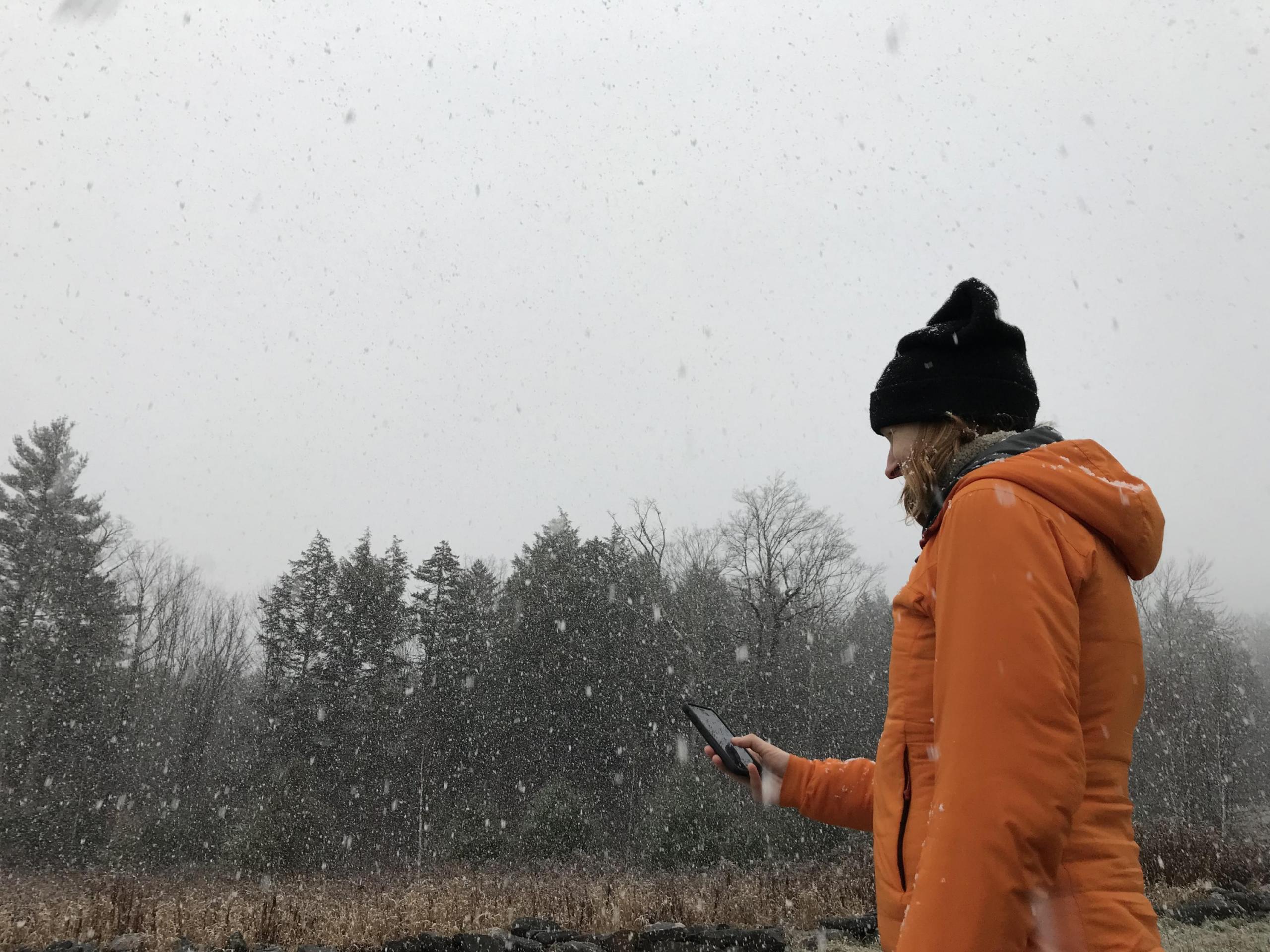 Photograph of a woman wearing an orange jacket and dark hat standing in a field with trees in the distance while she collects falling snow.